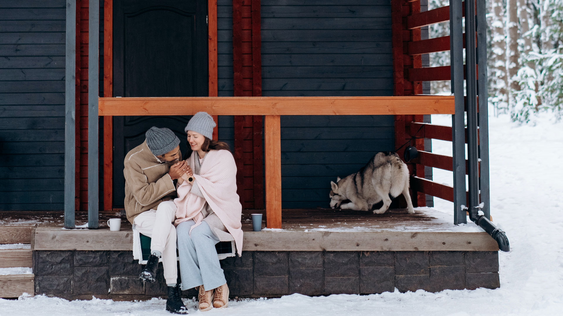 A couple, sitting together on their front porch in the winter, their dog playing behind them.