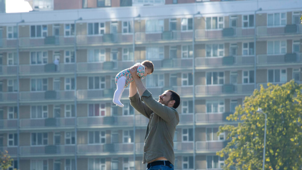 A man holding his daughter up and smiling at her, in front of their apartment building.
